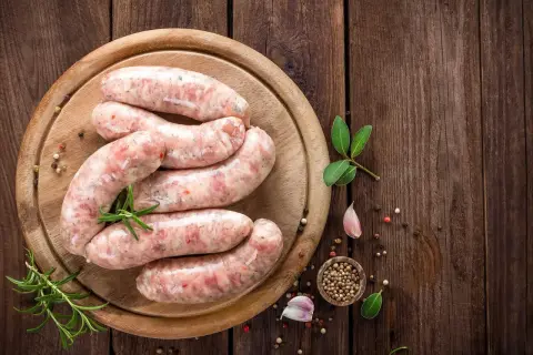 Surowa biała kiełbasa na desce, z ziołami i przyprawami. Zastanawiasz się, jak długo parzyć kiełbasę białą?