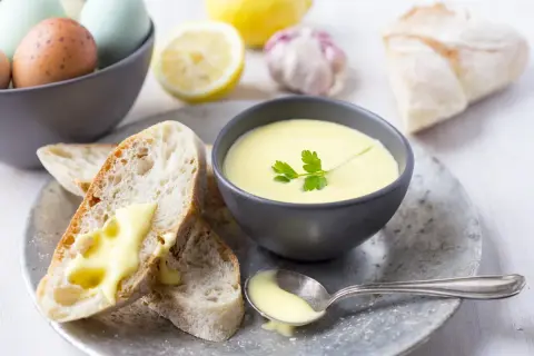 Kremowy sos aioli w miseczce, z pieczywem, jajkami, cytryną i czosnkiem w tle.