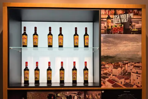 Butelki likieru Amaro Lucano ustawione na półkach, obok opisu produktu i włoskiego krajobrazu.