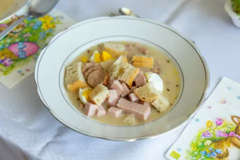 Biała zupa z kiełbasą, jajkiem i chlebem w talerzu, obok serwetki z zajączkiem.