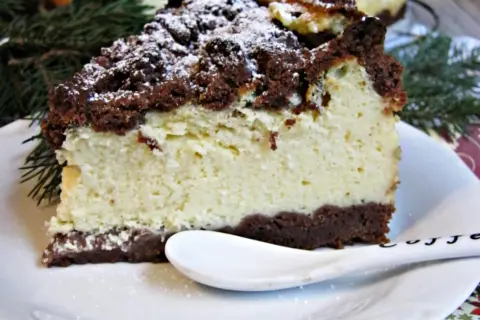 Kawałek puszystego sernika królewskiego z czekoladową kruszonką, posypany cukrem pudrem, na białym talerzyku z łyżeczką.