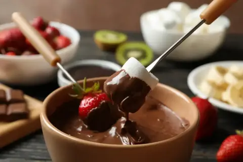 Czekoladowe fondue z pianką i truskawką na patyczku, w tle owoce i pianki.