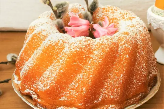 Wilgotna babka jogurtowa posypana cukrem pudrem, ozdobiona baziami i różowymi kwiatami.