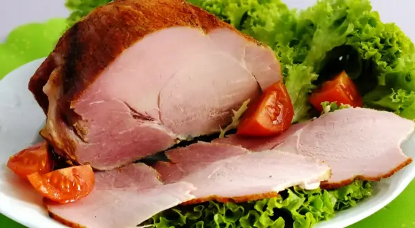 Pieczona szynka peklowana z sałatą i pomidorkami na białym talerzu.