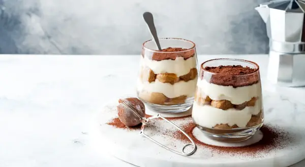 Dwa deserki tiramisu w szklankach, posypane kakao, na marmurowej desce.