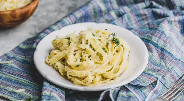 Talerz makaronu tagliatelle z kremowym sosem, posypany startym parmezanem i ziołami, na kolorowym obrusie.