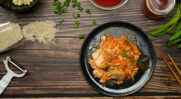 Talerz z kimchi, obok składniki: sezam, czosnek, papryka, szczypiorek, obieraczka i pałeczki.