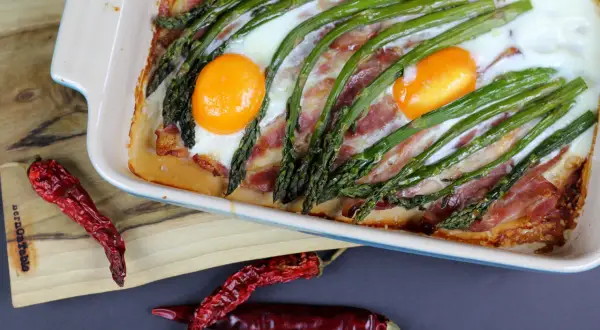 Zapiekane szparagi z boczkiem i jajkami w naczyniu, obok suszone papryczki chili.