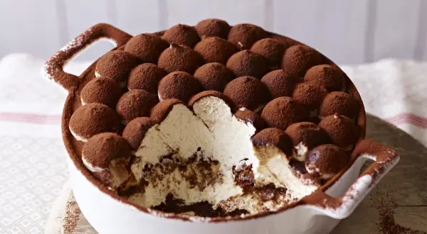 Tiramisu z mascarpone w białej misie, posypane kakao, z widocznym kremem i biszkoptami.