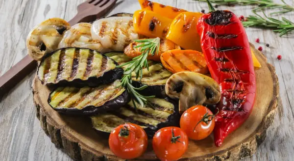Grillowane warzywa: bakłażan, papryka, pieczarki, pomidorki i marchewka na drewnianej desce.