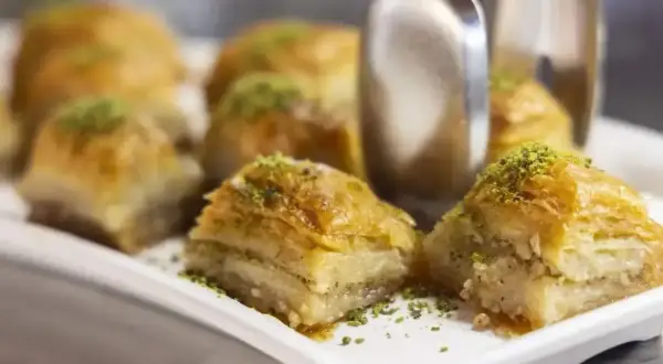 Słodka baklava z pistacjami na białej tacy, z widocznymi szczypcami do serwowania.