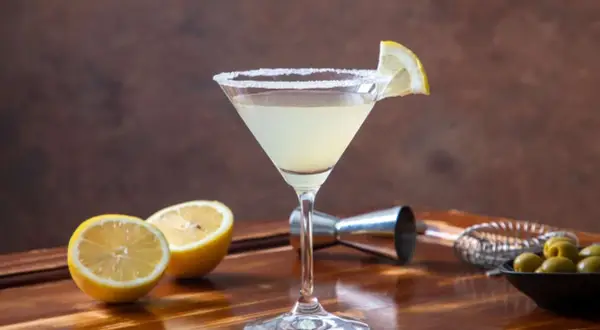 Koktajl Martini Bianco w kieliszku z solą na brzegu, ozdobiony plasterkiem cytryny, obok połówki cytryny i oliwek.