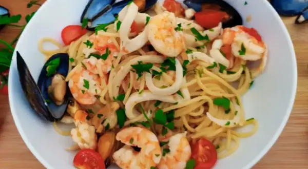 Talerz spaghetti z owocami morza: krewetkami, małżami i kalmarami, ozdobiony pomidorkami i natką pietruszki.