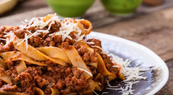 Talerz makaronu pappardelle z sosem bolognese i startym parmezanem.