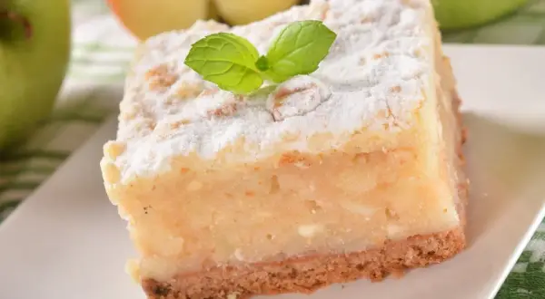 Kawałek ciasta z jabłkami, posypany cukrem pudrem i udekorowany listkiem mięty.