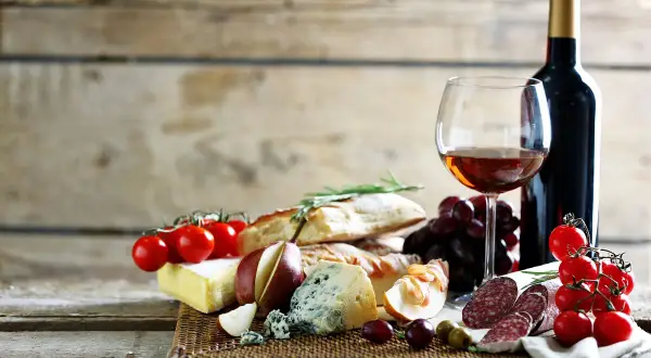 Butelka wina, kieliszek z winem, sery, pieczywo, winogrona, pomidorki i wędlina.