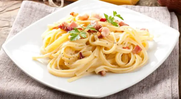 Porcja spaghetti carbonara z boczkiem i natką pietruszki na białym talerzu.