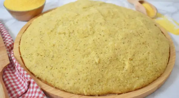 Gotowa, puszysta polenta na drewnianej desce, obok miseczka z mąką kukurydzianą.