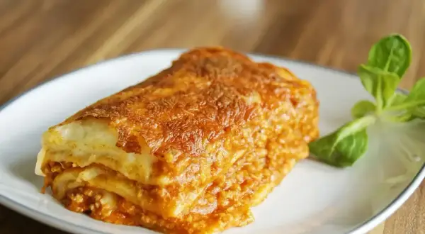 Kawałek zapieczonej lasagne z mięsem mielonym na białym talerzu, udekorowany listkami sałaty.