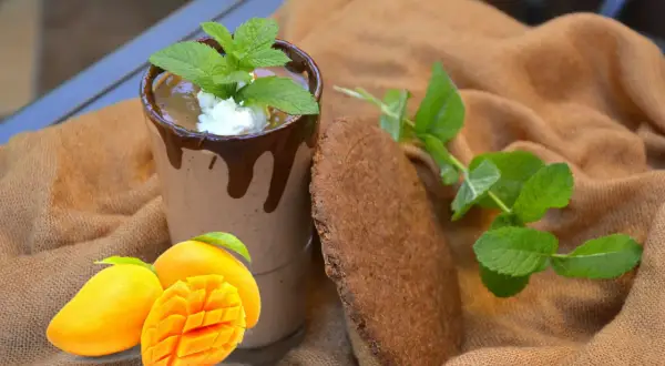 Szklanka mango lassi z czekoladą, miętą, mango i ciasteczkiem na brązowym tle.