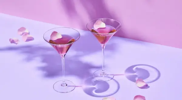 Dwa kieliszki z różowym drinkiem i płatkami róż na fioletowym tle.