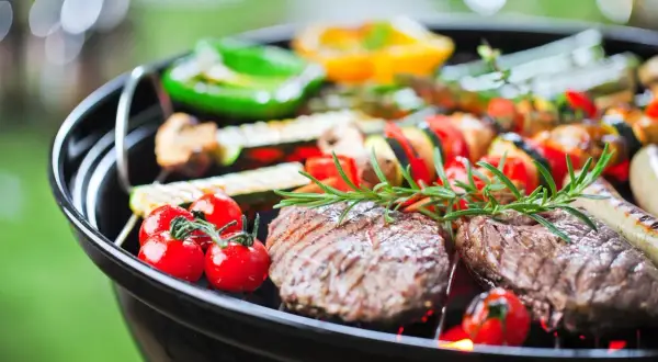 Grill z mięsem, warzywami i ziołami na tle zieleni.