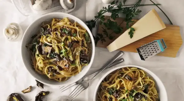 Dwa talerze makaronu z grzybami, posypane parmezanem i natką pietruszki. Obok świeże i suszone grzyby, kawałek parmezanu i tarka. Pyszny makaron z oliwą truflową.