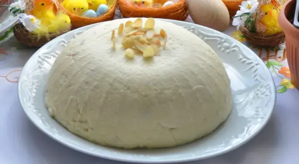 Pascha na białym talerzu, ozdobiona płatkami migdałów, w tle wielkanocne kurczaczki.