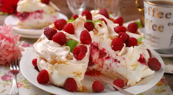 Tort bezowy z bitą śmietaną, malinami i listkami mięty na białym talerzu.