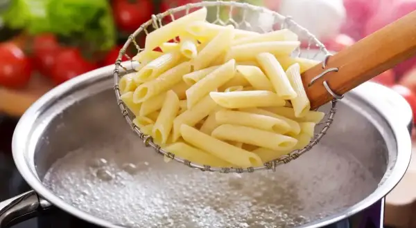 Makaron penne w cedzaku nad garnkiem z gotującą się wodą.