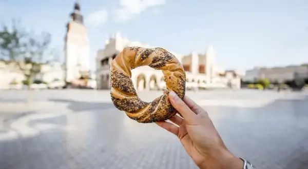 Dłoń trzyma obwarzanka krakowskiego z makiem na tle Sukiennic.