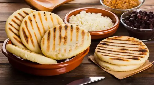 Grillowane arepy w misce, obok miseczki z białym serem, czarną fasolą i brązowym gulaszem.