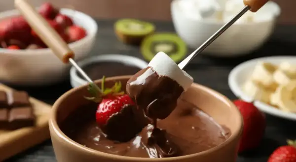 Czekoladowe fondue z pianką i truskawką na patyczku, w tle owoce i pianki.