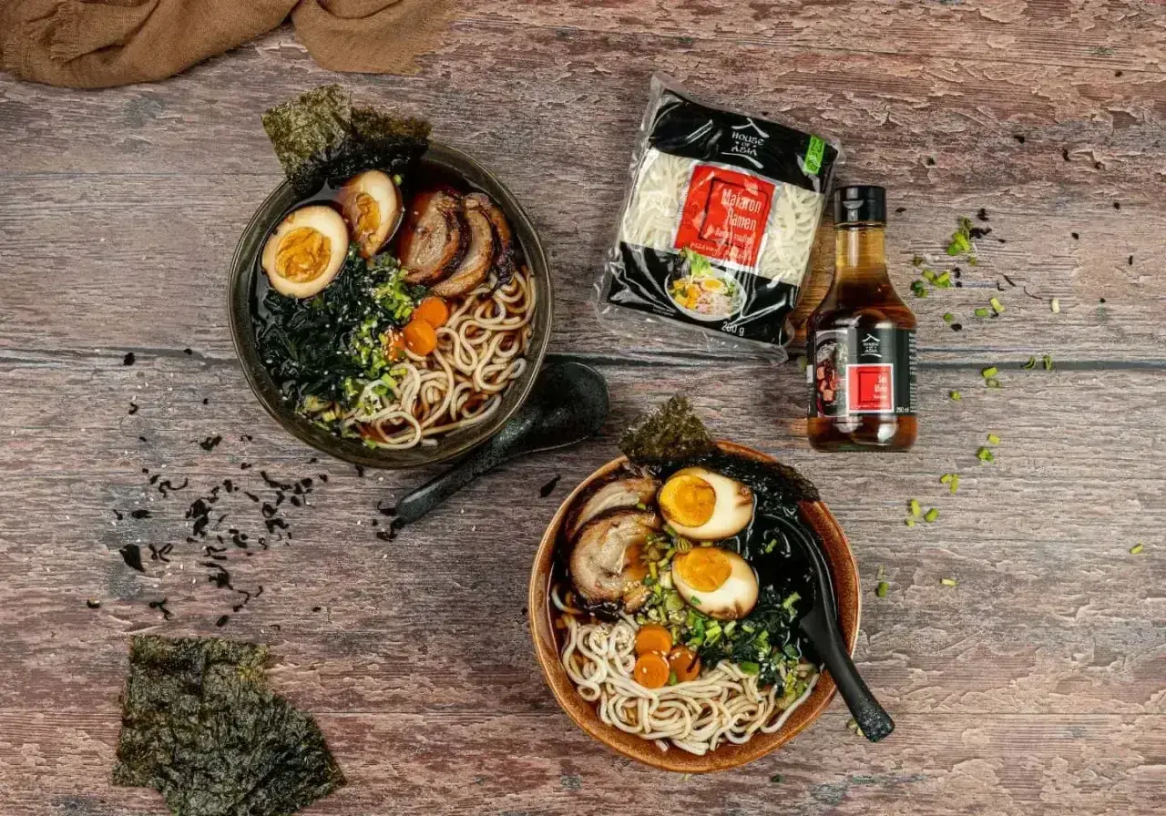 Dwie miski domowego ramenu z jajkiem, mięsem i nori, obok makaron i sos.