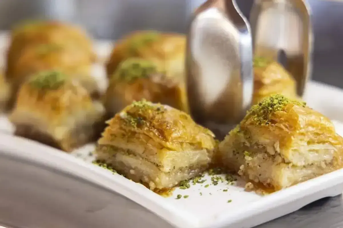Słodka baklava z pistacjami na białej tacy, z widocznymi szczypcami do serwowania.