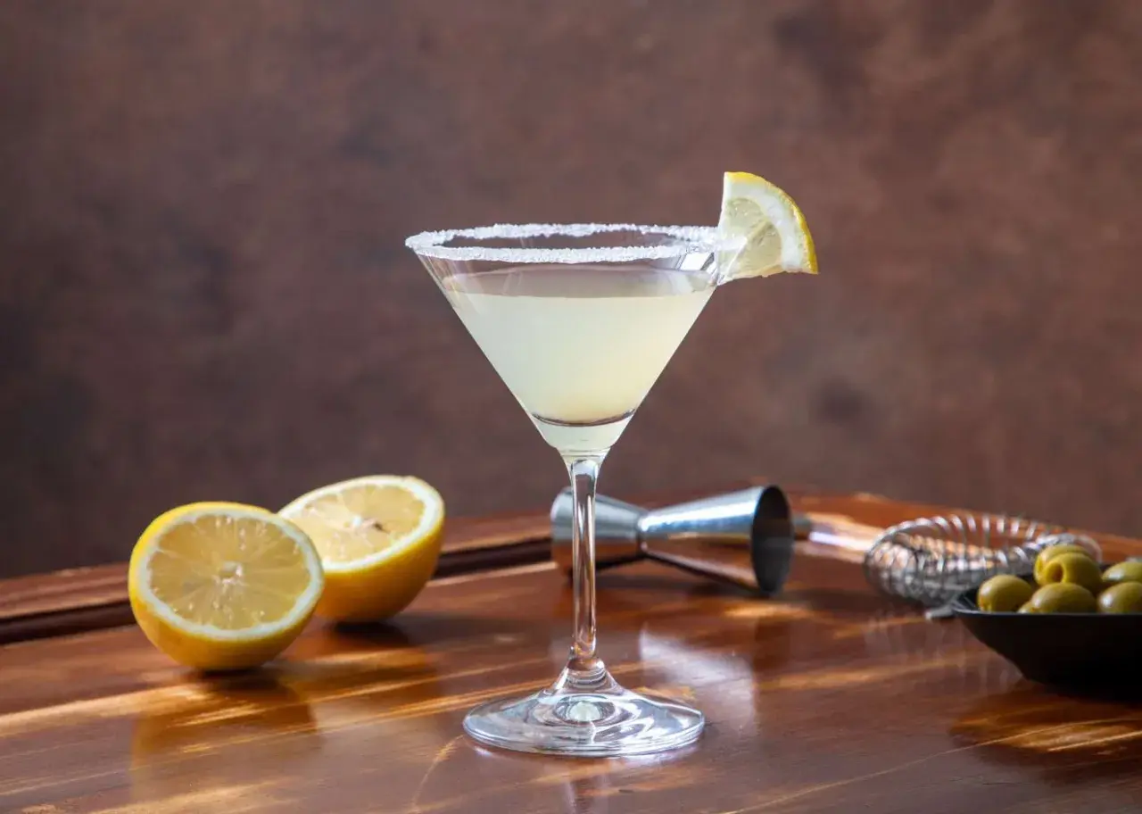 Koktajl Martini Bianco w kieliszku z solą na brzegu, ozdobiony plasterkiem cytryny, obok połówki cytryny i oliwek.