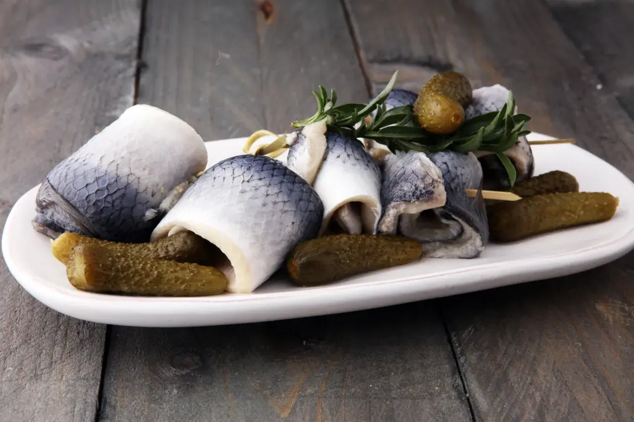 Roladki ze śledzia z ogórkami konserwowymi i gałązką rozmarynu na białym talerzu.