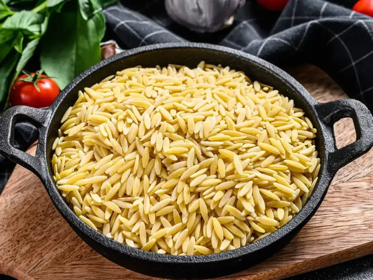 Makaron orzo w czarnej patelni, obok pomidorów i ziół.