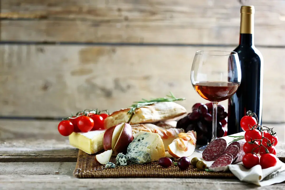 Butelka wina, kieliszek z winem, sery, pieczywo, winogrona, pomidorki i wędlina.