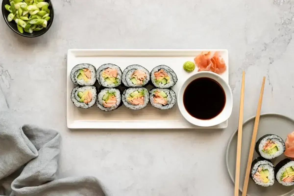 Ułożone na talerzu cienkie rolki sushi z łososiem i awokado, obok sos sojowy, wasabi i imbir.