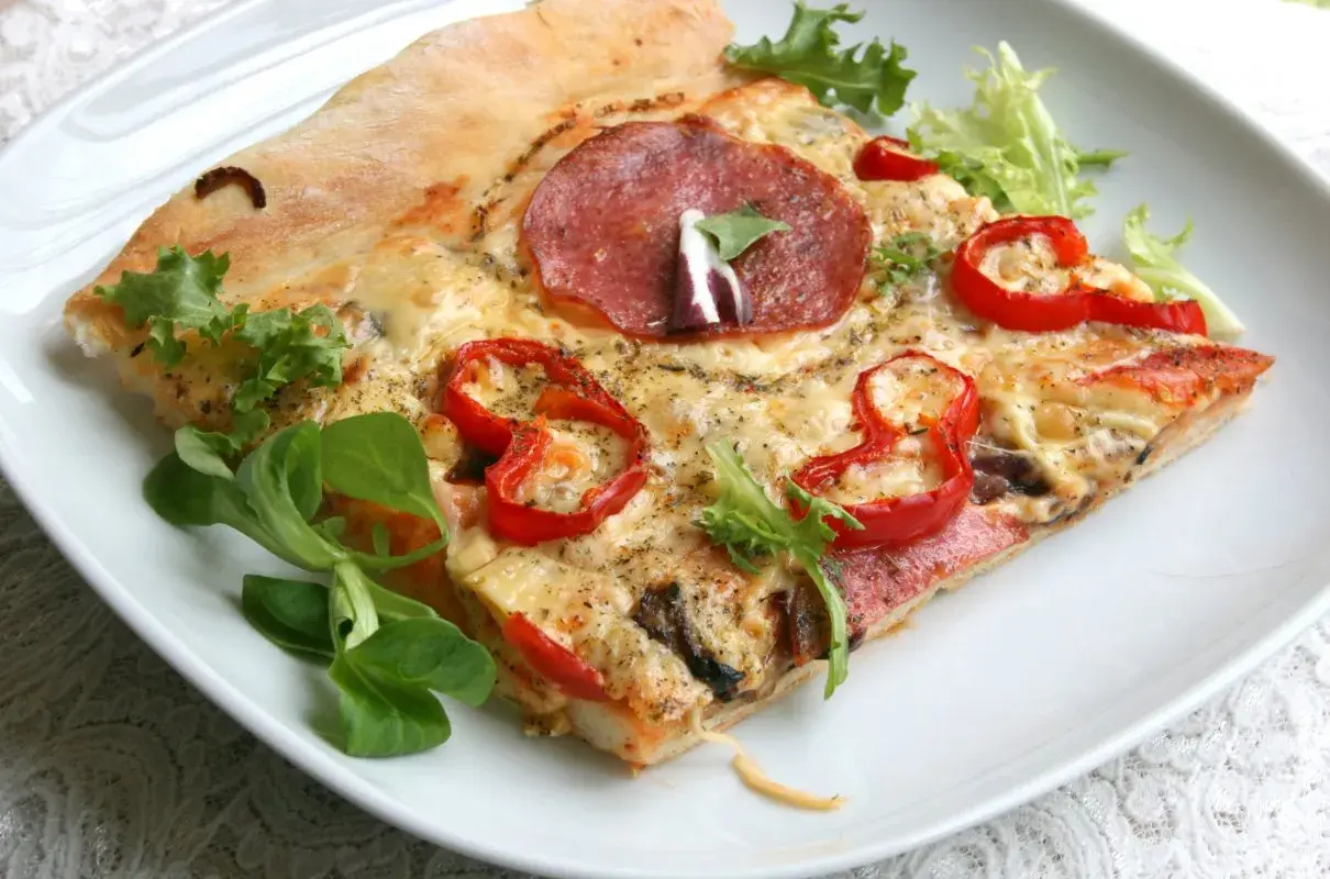 Kawałek pizzy z salami, papryką i serem na białym talerzu, ozdobiony sałatą.