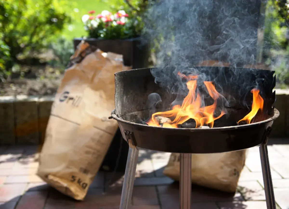 Płonący grill z workami węgla drzewnego w tle.