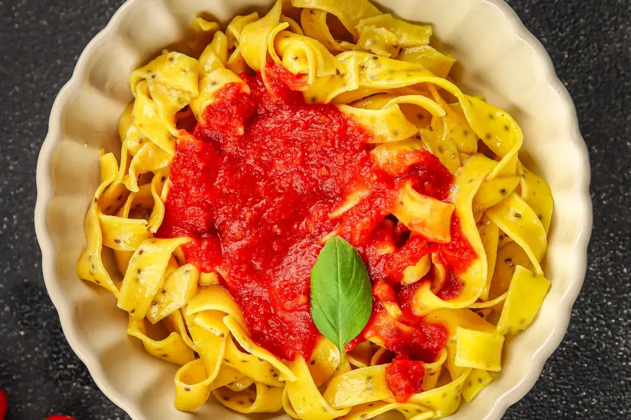Szeroki makaron tagliatelle z sosem pomidorowym i listkiem bazylii w białej misce.