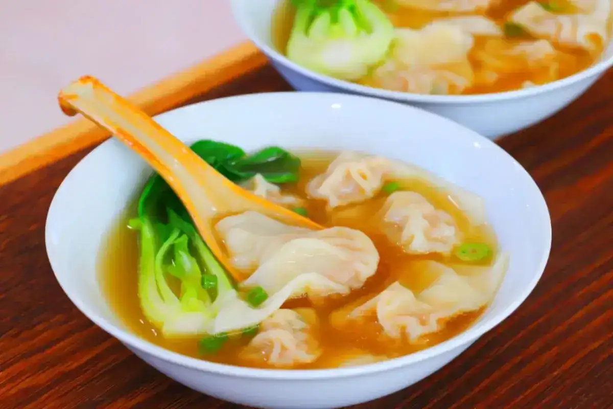 Dwie białe miski z zupą wonton, z widocznymi pierożkami, zielonymi warzywami i pomarańczową łyżką.