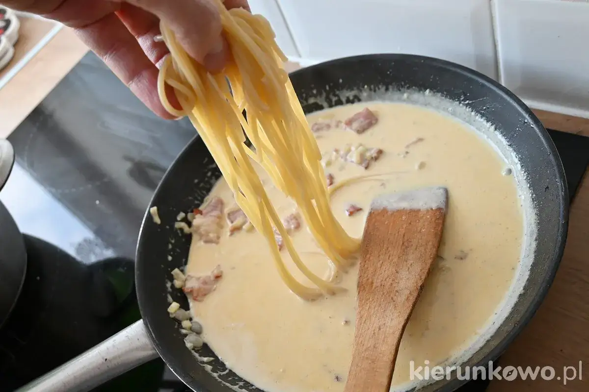 Dłoń dodaje spaghetti do sosu carbonara na patelni.