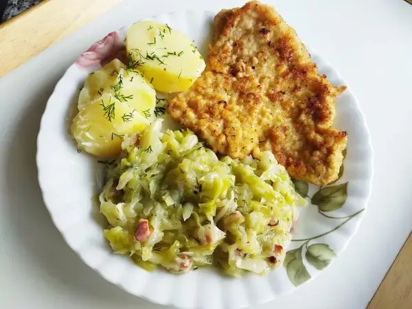 Kotlet schabowy z ziemniakami i zasmażaną kapustą z boczkiem na talerzu.
