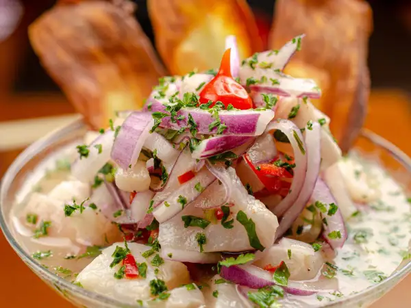 Ceviche z ryby, cebuli, papryczki chili i kolendry w szklanej misce, z chipsami bananowymi w tle.