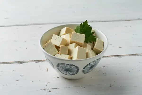 Miseczka z pokrojonym w kostkę tofu i listkiem pietruszki na białym, drewnianym tle.