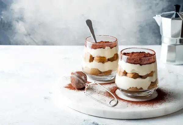 Dwa deserki tiramisu w szklankach, posypane kakao, na marmurowej desce.