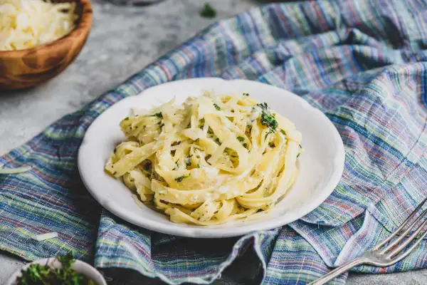 Talerz makaronu tagliatelle z kremowym sosem, posypany startym parmezanem i ziołami, na kolorowym obrusie.
